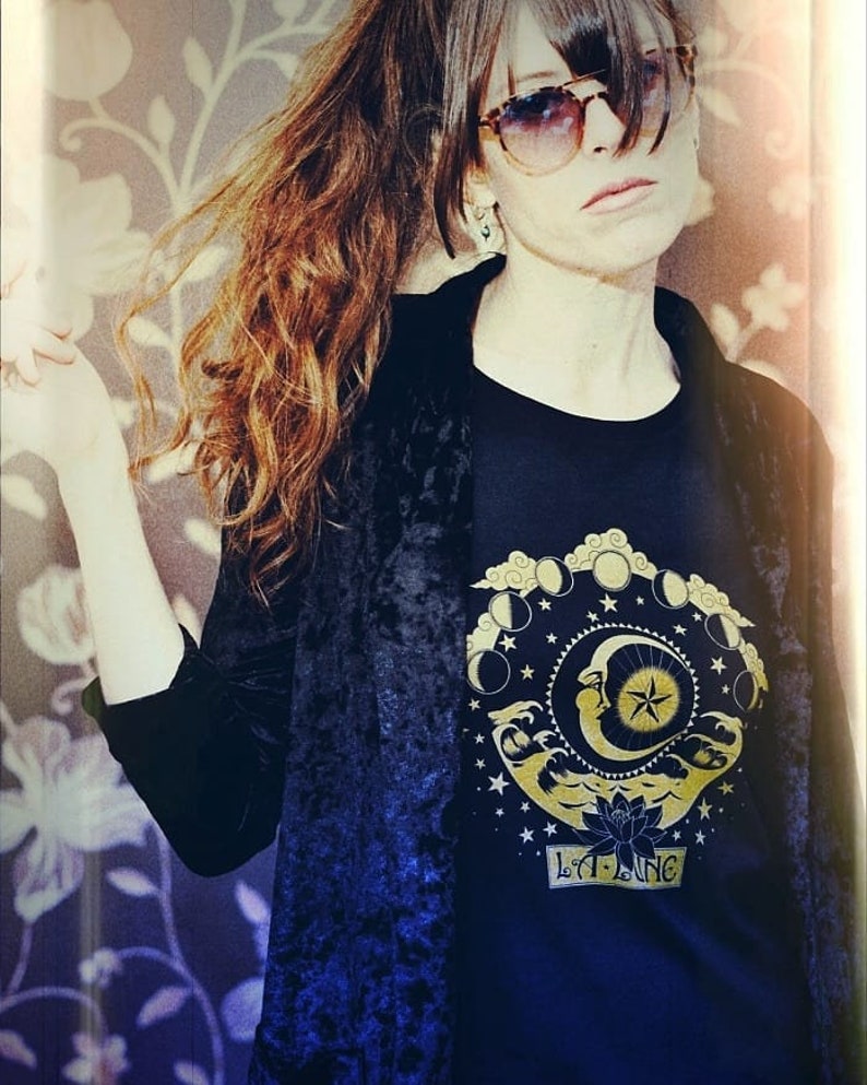 May include: A woman wearing a black velvet jacket over a black t-shirt with a gold moon and stars design. The t-shirt also has the text "LILA LANG" in gold lettering.