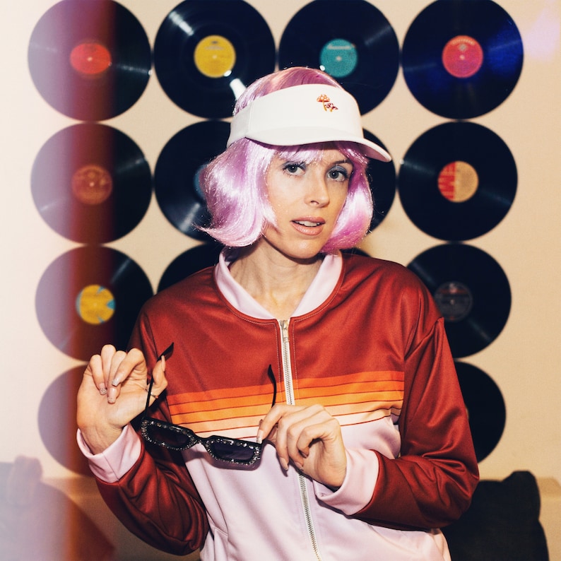 May include: A woman with pink hair is wearing a white visor with a mushroom graphic and a red and pink striped jacket. She is holding a pair of black sunglasses.