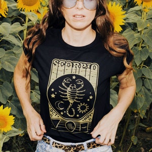 May include: A black t-shirt with a gold scorpion graphic and the text "Scorpio" with dates "Oct 23 - Nov 21". The shirt is worn by a person in a field of sunflowers.