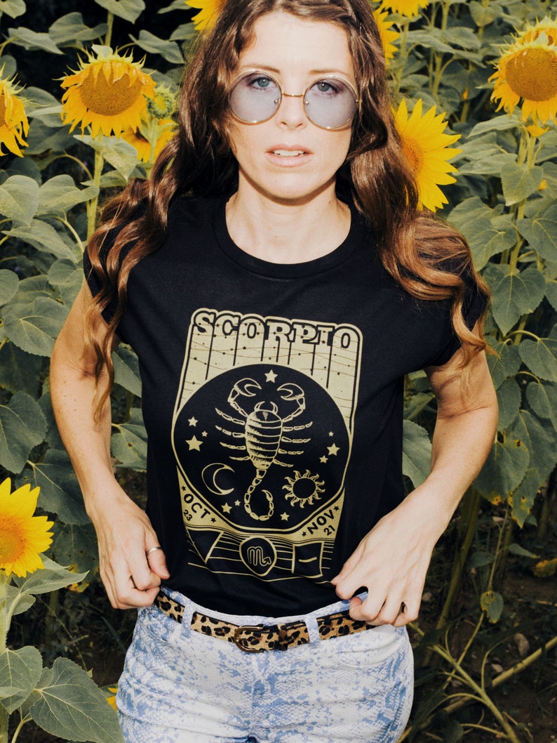 May include: A black t-shirt with a gold scorpion graphic and the text "Scorpio" with dates "Oct 23 - Nov 21". The shirt is worn by a person in a field of sunflowers.