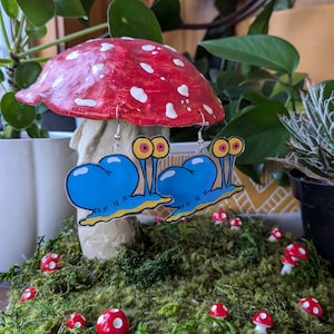 May include: A pair of blue snail earrings with yellow eyes and a white shell, displayed on a red and white mushroom with green moss.