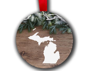May include: A round, white, wooden ornament with a white outline of the state of Michigan. The ornament is decorated with a green pine garland with white snow on the branches. The text "SCENE HAPPY" is printed on the bottom of the ornament.