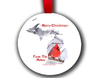 May include: A white round Christmas ornament with a red cardinal bird perched on a branch in a snowy Michigan outline. The ornament has the text "Merry Christmas" and "From The -Mitten- SCENE HAPPY" printed on it.