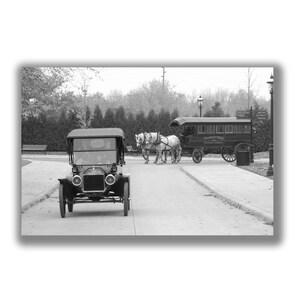 May include: A black and white photo of a vintage car driving down a paved road. The car is a Model T Ford with a black canvas top. The car is driving past a horse-drawn carriage and a vintage bus. The bus is labeled "Greenfield Village" and "Henry Ford Museum".