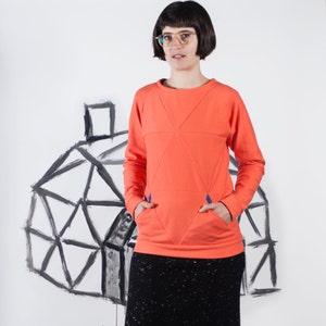 May include: A woman wearing a bright orange sweatshirt with black pockets and a black skirt stands in front of a white backdrop with a drawing of a geodesic dome. She is wearing glasses and has short dark hair.