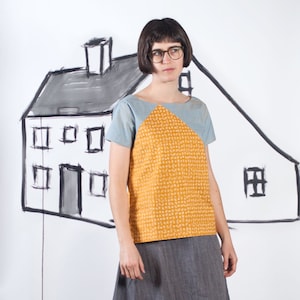 May include: A woman wearing a two-tone top with a yellow patterned front and a gray skirt. The top has a light blue sleeve and shoulder. The background features a large drawing of a house. The setting appears to be a studio with sewing equipment.