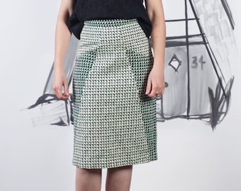 May include: A woman wearing black-framed glasses and a black top with a green and white checkered skirt. She is standing in front of a white backdrop with a black and white drawing of a house.