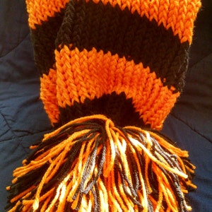 May include: A knitted scarf in black and orange stripes with a matching tassel. The scarf is made of thick yarn, with alternating bands of black and orange. The tassel at the end is a mix of black and orange yarn.