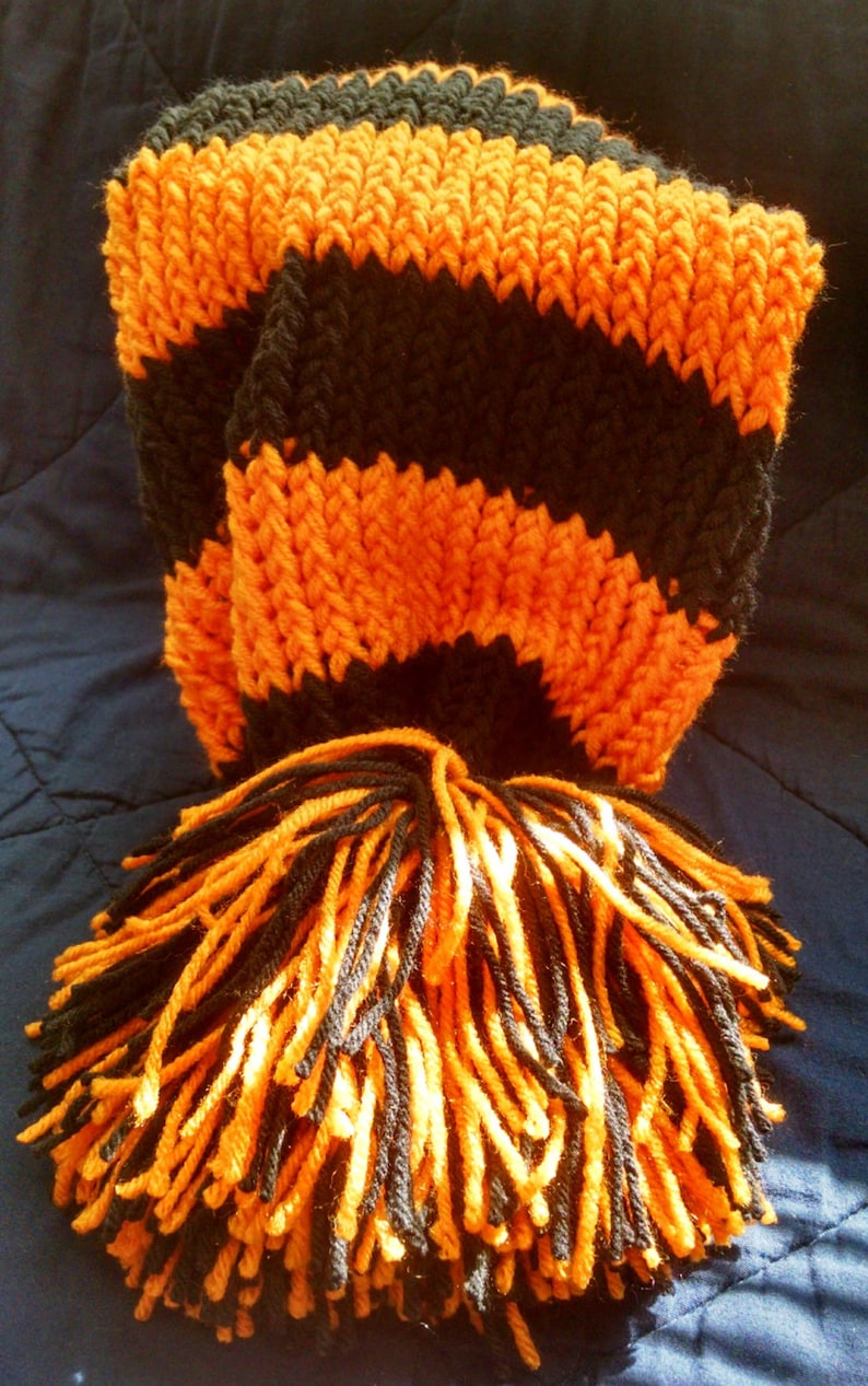 May include: A knitted scarf in black and orange stripes with a matching tassel. The scarf is made of thick yarn, with alternating bands of black and orange. The tassel at the end is a mix of black and orange yarn.