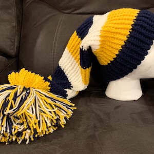May include: A long, striped knitted hat in navy blue, yellow, and white, with a large tassel. The hat is displayed on a white mannequin head. The tassel is a mix of navy blue, yellow, and white yarn.