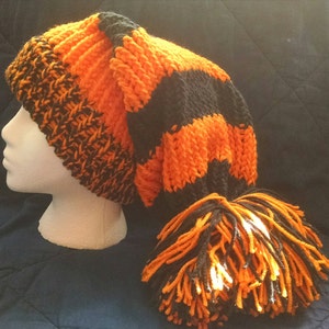 May include: A hand-knitted orange and black hat with a long, pointed shape and a tassel. The hat is displayed on a white mannequin head. The hat's design features alternating stripes of orange and black.