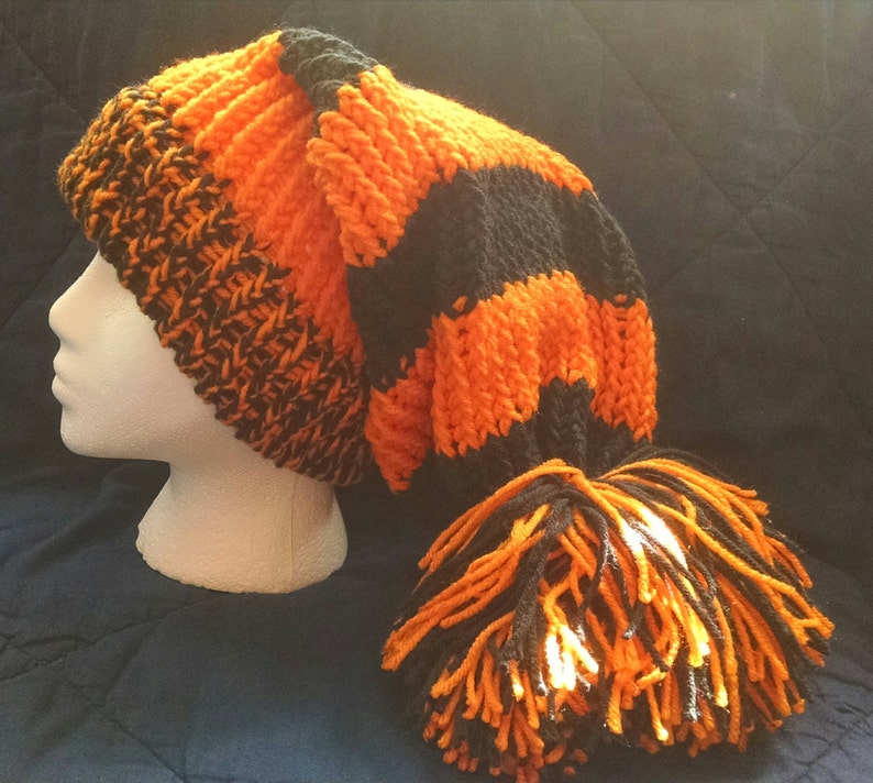 May include: A hand-knitted orange and black hat with a long, pointed shape and a tassel. The hat is displayed on a white mannequin head. The hat's design features alternating stripes of orange and black.