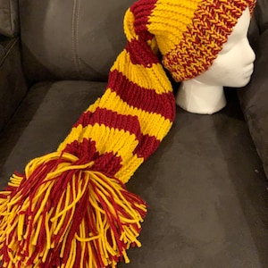 May include: A long, red and yellow striped knitted hat with a pom-pom. The hat is made of yarn and has a long, flowing scarf attached to it.