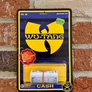 Puede incluir: Un paquete de juguetes novedoso con el logotipo de Wu-Tang Clan en amarillo sobre un fondo azul y negro. El paquete incluye un fajo de dinero de juguete sujeto con una banda roja, y el texto "CASH RULES EVERYTHING AROUND ME".