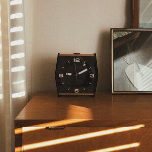 May include: A black alarm clock with gold accents sits on a wooden dresser with a drawer. The clock face has black numbers and hands, and the clock is set to 10:10. The dresser is made of wood and has a light brown finish. The dresser is in a room with a white wall and a window with white blinds.