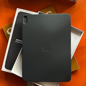 May include: A black iPad Air Smart Folio case with the Apple logo. The case is displayed on top of a white box and a book. The background is orange.