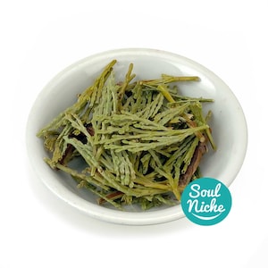 May include: A small, white, oval-shaped bowl filled with dried, green, needle-like herbs. The herbs are a light green color and appear to be dried. The bowl is set against a white background. The text "Soul Niche" is in a teal circle.