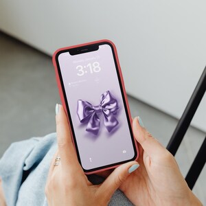 Op de afbeelding: Een roze telefoonhoesje met een paarse strik op het scherm. Het vergrendelscherm van de telefoon toont de tijd "3:18" en de datum "Maandag 13 januari".