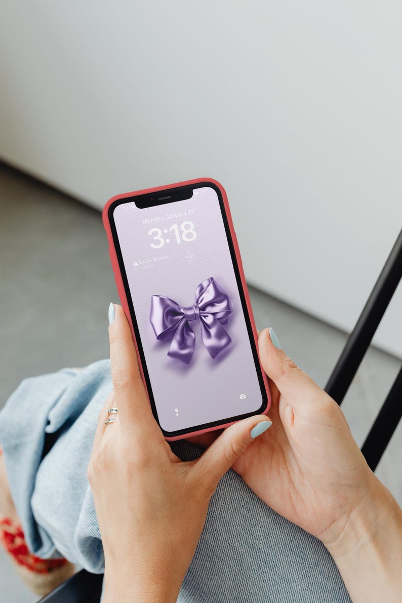 Op de afbeelding: Een roze telefoonhoesje met een paarse strik op het scherm. Het vergrendelscherm van de telefoon toont de tijd "3:18" en de datum "Maandag 13 januari".