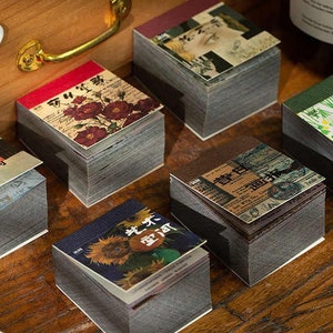 May include: A collection of small, decorative notepads with vintage-style designs. Each notepad features a unique cover with floral, botanical, or artistic imagery. The notepads are stacked on a wooden surface, creating a visually appealing arrangement.