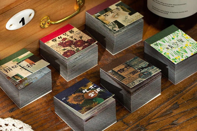 May include: A collection of small, decorative notepads with vintage-style designs. Each notepad features a unique cover with floral, botanical, or artistic imagery. The notepads are stacked on a wooden surface, creating a visually appealing arrangement.