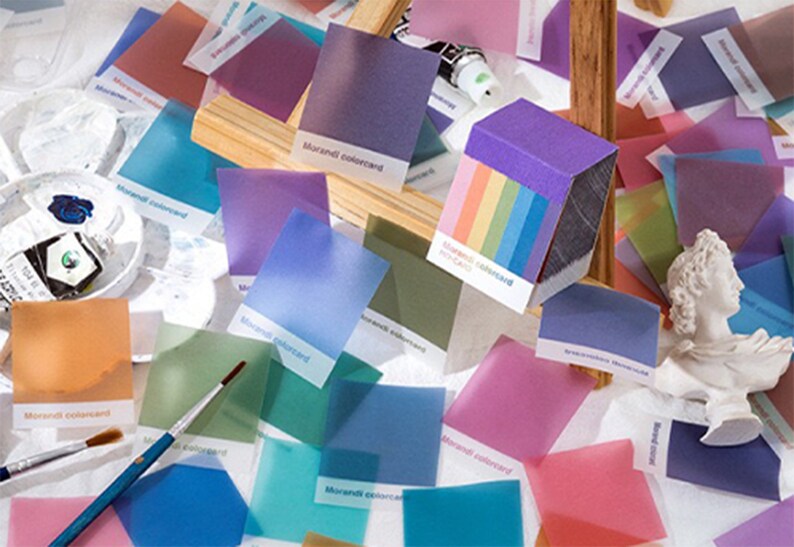 May include: A collection of Morandi color cards in various pastel shades, including pink, blue, and purple. The cards are scattered around art supplies like paint tubes, brushes, and a small bust. The text "Morandi colorcard" is visible on some cards.