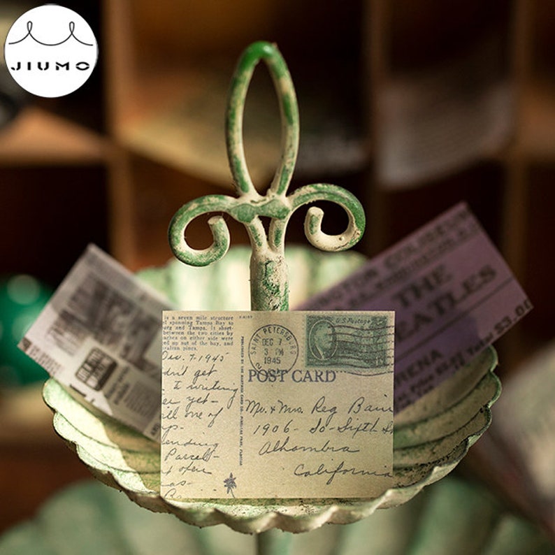 May include: A vintage-style metal card holder with a distressed green finish. It holds a postcard with handwritten text and a stamp, along with a ticket stub. The postcard reads "POST CARD" and "California".