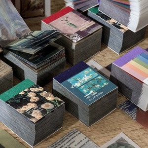 May include: Stacks of decorative paper cards with various designs, including floral, artistic, and text-based imagery. The cards have a gray edge and are arranged on a wooden surface. Some cards feature the text "MO-CARD".
