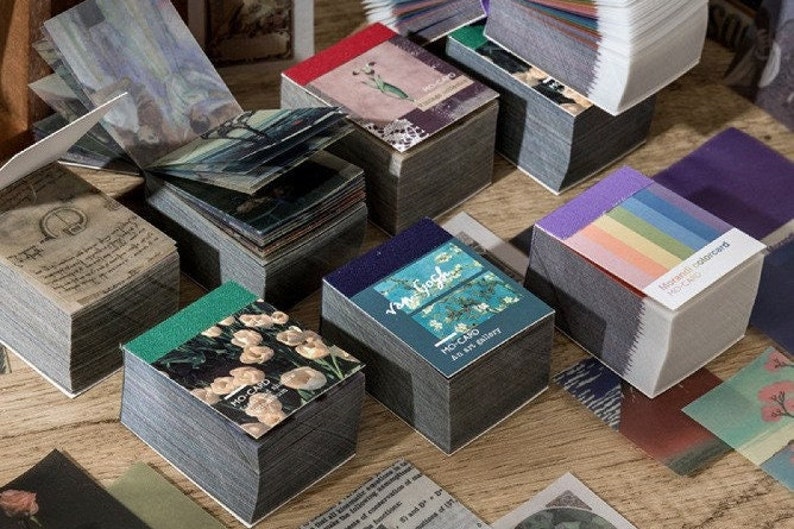 May include: Stacks of decorative paper cards with various designs, including floral, artistic, and text-based imagery. The cards have a gray edge and are arranged on a wooden surface. Some cards feature the text "MO-CARD".