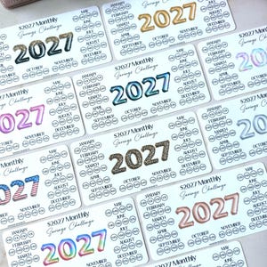 May include: A collection of rectangular white paper savings challenge trackers for the year 2027. Each card features the text $2027 Monthly Savings Challenge, the year 2027 in various patterns, and circular icons labeled with months and dollar amounts.