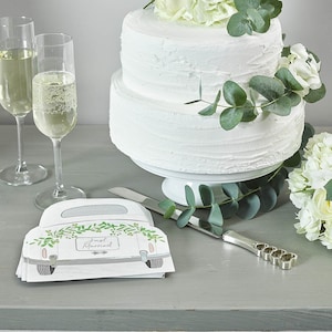 May include: A two-tiered white wedding cake decorated with white flowers and greenery. The cake is on a white cake stand. There are two champagne flutes and a cake knife on the table. There are also napkins with a car design and the text "Just Married" on them.