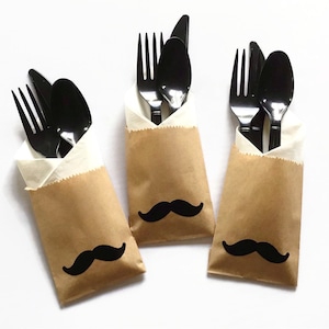 May include: Three brown paper bags with black paper napkins inside. Each bag has a black paper moustache sticker on the front. Each bag contains a set of black plastic cutlery, including a fork, knife, and spoon.