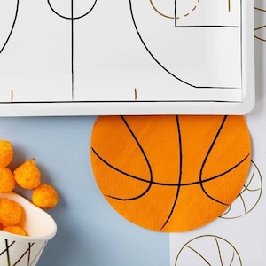 May include: Orange and black basketball-themed paper napkin with a basketball design. The napkin is on a blue background with other basketball-themed napkins.