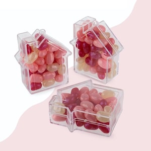 May include: Three clear, house-shaped containers filled with colorful jelly beans. The jelly beans are in shades of pink, red, and yellow. The containers are arranged on a light pink background.