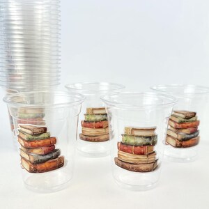 May include: Clear plastic cups with a stack of books design. A stack of clear cups is in the background. Four cups with book graphics are in the foreground, with a variety of book colors.