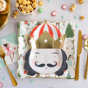May include: A Nutcracker themed party table setting with gold cutlery, a green and white napkin, a white paper plate with a Nutcracker face, and a bowl of popcorn.