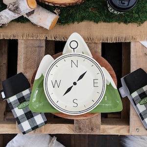 May include: Green and white paper plates shaped like a compass with a mountain design. The plates feature the words "N", "S", "E", and "W" on the compass face.