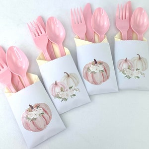 May include: Four pink plastic forks, spoons, and knives are wrapped in white paper bags with a pink and white pumpkin design. The bags are decorated with a watercolour-style pumpkin with white flowers.