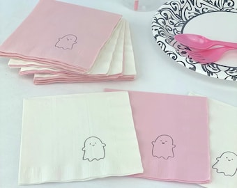 May include: Pink and white paper napkins with a ghost design. The ghost has a smiling face and is outlined in black. The napkins are stacked on a white surface.