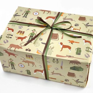 Może przedstawiać: Brązowe i zielone pudełko prezentowe z papierem do pakowania w stylu myśliwskim. Papier zawiera ilustracje ps&oacute;w, jeleni, kompasu, karabinu i innych akcesori&oacute;w myśliwskich.