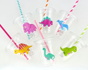 May include: Six clear plastic cups with colorful striped straws and dinosaur stickers. The dinosaurs are pink, green, blue, and yellow.