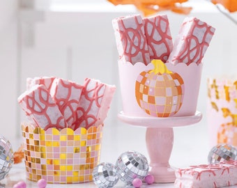 Puede incluir: Golosinas de malvavisco rosa y blanco con un diseño de espiral rosa y blanco, exhibidas en recipientes en forma de calabaza rosa y dorado con un diseño de bola de discoteca. Los recipientes están sobre una superficie blanca con bolas de discoteca plateadas y caramelos rosas.