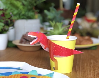 May include: A yellow paper cup with a red dinosaur design and a red and yellow striped straw. The dinosaur is holding the cup with its arms and legs. The cup is on a wooden table with a blue and orange dinosaur-themed paper plate.