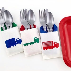 May include: Three white paper cutlery holders with truck silhouettes in blue, green, and red. Each holder contains a fork, spoon, and knife. A red rectangular plate is on the right.