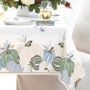 Pode incluir: Uma toalha de mesa branca com uma borda de abóboras em tons de verde, azul e bege. As abóboras são estilizadas e têm folhas e videiras. O texto "Rise and Shine" está escrito em uma taça de vinho.