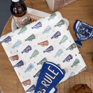 K&ouml;nnte beinhalten: Ein wei&szlig;er Pappteller mit einem goldenen und blauen Megaphon-Muster. Die Megaphone haben verschiedene Spr&uuml;che darauf, darunter "Dads Rule", "Best Dad", "You Rock", "Love You" und "No. 1 Dad". Es gibt einen blauen Megaphon-Ausschnitt mit dem Text "Dads Rule" darauf.