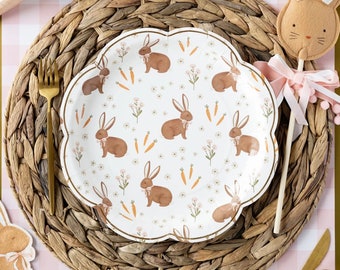 May include: White paper party plate with a scalloped edge and a repeating pattern of brown bunnies, carrots, and flowers. The plate is on a woven brown placemat.