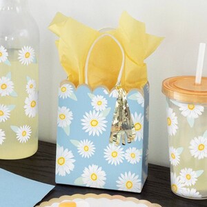 May include: A blue and white daisy patterned gift bag with a gold tassel. The bag is filled with yellow tissue paper and is sitting on a table with a clear glass pitcher and a clear plastic cup with a straw.
