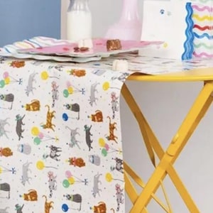 Könnte beinhalten: Ein weißer Tischläufer mit einem sich wiederholenden Muster von Katzen, die Partyhüte tragen und Ballons halten. Der Läufer liegt auf einem gelben Klapptisch mit einem blauen Wollknäuel auf dem Boden davor.