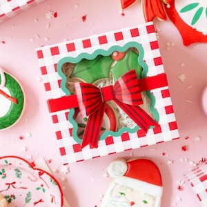 May include: Overhead shot of Christmas-themed cookies and gift boxes on a pink surface. Cookies include a wreath, ornament, bow, and Santa Claus. Gift boxes are red and white checkered with red bows. Festive holiday treats.